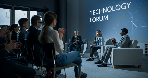 International Technology Conference: Host Asking Caucasian Female Tech CEO a Question In Front Of Audience. Successful Woman Delivering Inspirational Speech And Diverse Attendees Applaudding Her.