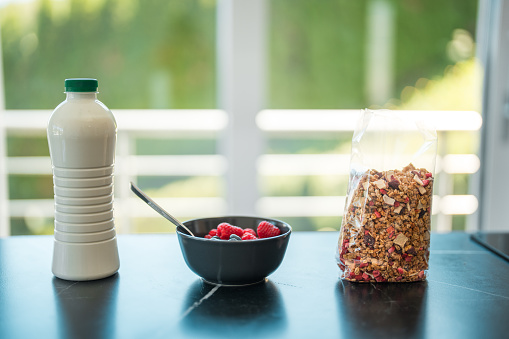 Image of a healthy breakfast prepared on a chic kitchen counter, bowl with various berries and a spoon in it, a bottle of milk and a bag of cereal next to it. Big window with daylight in the background.