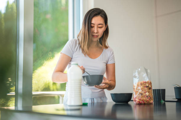 Waist up image of a young  Asian woman standing behind a kitchen surface  while holding a bowl and mixing  cereal in it in the early morning. Various bowls and a bottle of dairy  on the counter, pot on the stove and a cup of tea ready.