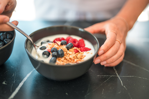 Close up of a cereal mix  in low-fat yogurt  and various berries in a chic ceramic bowl, another bowl with only blueberries in, next to the first one. A hand holding a small spoon and starting to to dig in, the other hand holding the bowl still on the table. High angle shot.