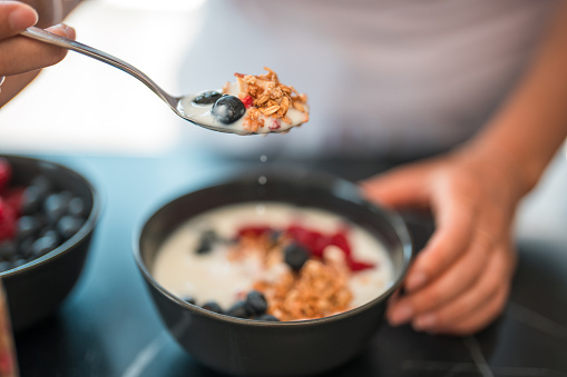 Close up of a spoon holding  cereal in low-fat yogurt  and blueberries above the black  ceramic bowl on the kitchen counter, another bowl with only blueberries in, next to the first one. The other hand holding the bowl still from the side. High angle shot.