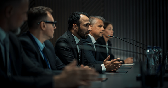 Middle Eastern Male Head Of Political Delegation Speaking At International Economic Forum Press Conference. High-Profile Politician Reporting On Successful Agreements. Diverse Delegates Listening.