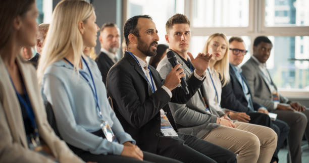 Multiethnic Male Technology Conference Attendee Sittign In Crowd And Using Microphone To Ask A Question Related To Presentation Of An Innovative Gadget. Middle Eastern Man At International Tech Forum. Multiethnic Male Technology Conference Attendee Sittign In Crowd And Using Microphone To Ask A Question Related To Presentation Of An Innovative Gadget. Middle Eastern Man At International Tech Forum. business conference stock pictures, royalty-free photos & images