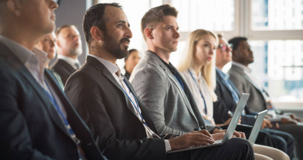 istock Multiethnic Man Sitting in a Crowded Auditorium at Conference. Middle Eastern Businessman Listening To Keynote, Typing On Laptop. Specialist Watching Technology Presentation About Software Solutions. 1757748559