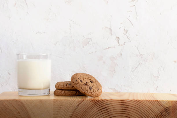 Glass of milk and cookies on wooden serving board, white textured wall as background. Copy space.