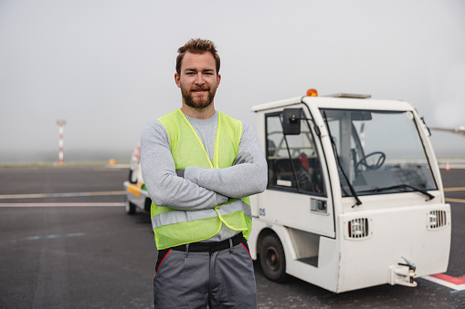 Portrait of a confident young adult, standing in his reflective jacket. He is working as part of the airport ground crew.