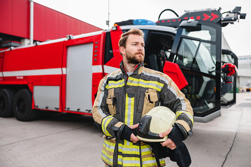 A young firefighter posing in uniform and helmet in hand inside the fire station.