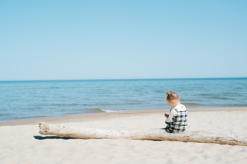 Lonely child sitting on a log on the beach by the sea, copy space. Rear view of a little caucasian girl playing alone on the beach on a sunny summer day.