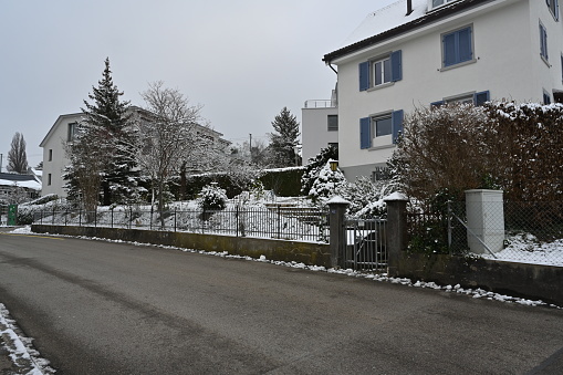 Urdorf, Switzerland- 12 18 2022: Street in residential quarter of village Urdorf in Switzerland in winter. There is a thin layer of snow especially on threes. Two incidental persons are walking on the background.