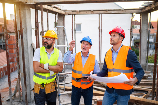 A group of male Caucasian architect, building contractor, and engineer, having a meeting on a construction site, where they analyze the blueprint and how the project is going