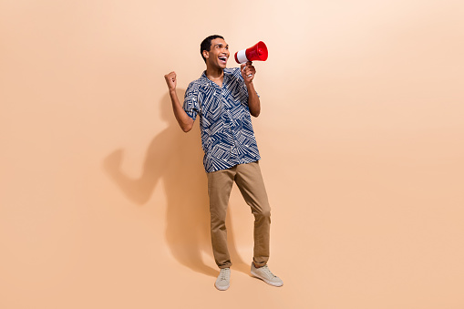 Full length photo of excited ecstatic man anouncing sale in megaphone look empty space clenching fist isolated on beige color background.