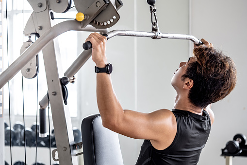 Asian sport man with well trained body in black sportswear pulling overhead bar doing exercise training with lat pulldown machine in fitness gym. Muscular build and physical training concept