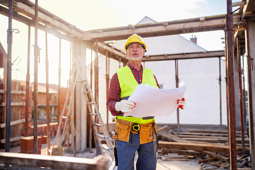 Senior Caucasian male architect holding an blueprint, while observing the work on a construction site