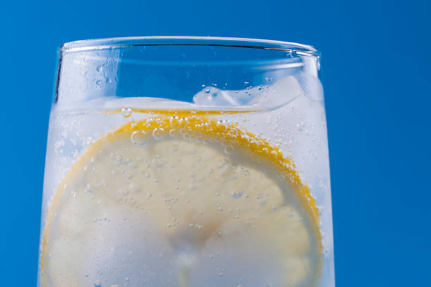 Sparkling water Summer fresh drink - cold sparkling water in a glass with some ice and slice of a lemon over a solid blue background. soda water glass lemon stock pictures, royalty-free photos & images