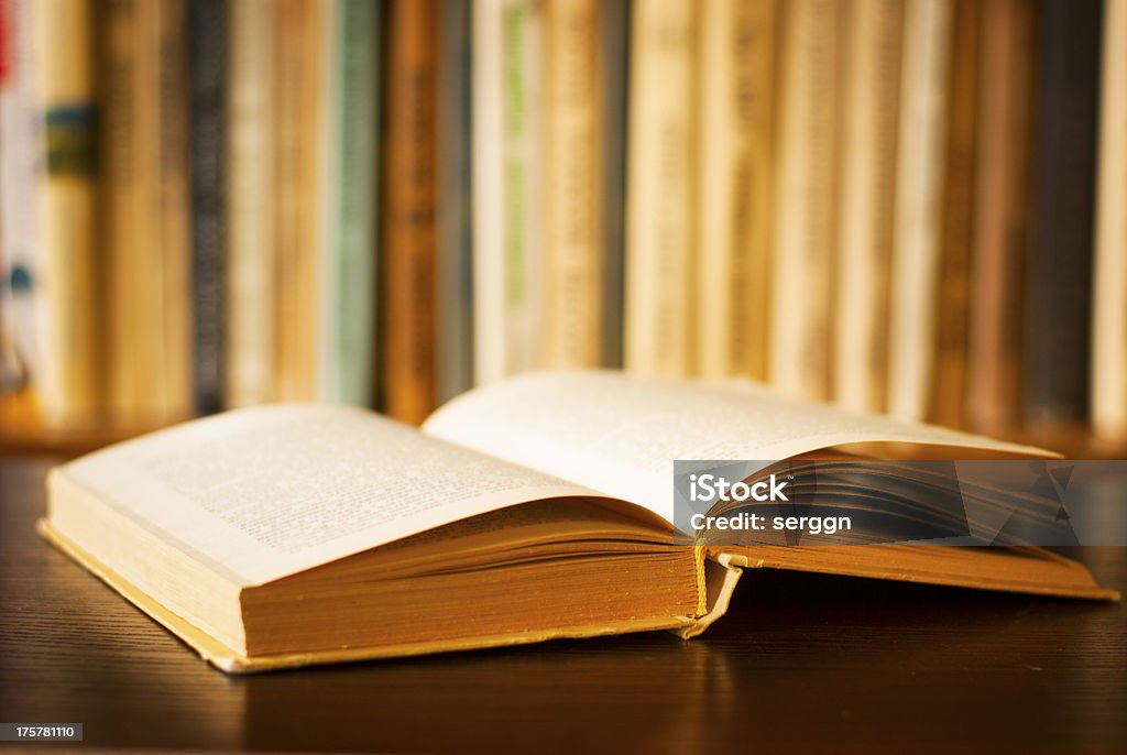 Open book Low angle view of a large open hardcover book lying on a wooden desk in front of a shelf of books with shallow dof Book Stock Photo