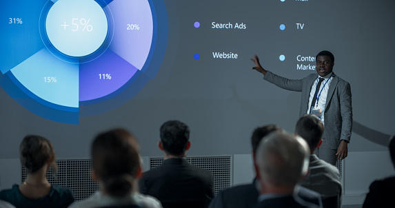 Corporate Event: Black Male Marketing Director Giving Presentation To Colleagues or Investors In Conference Room Of Innovative Startup Office. African Man Talking About Advertising Channels, Budget.