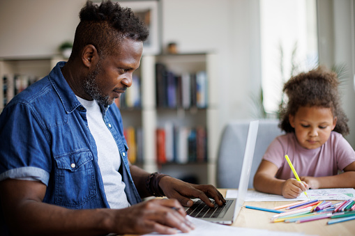 Father working on laptop while his daughter drawing beside him at home