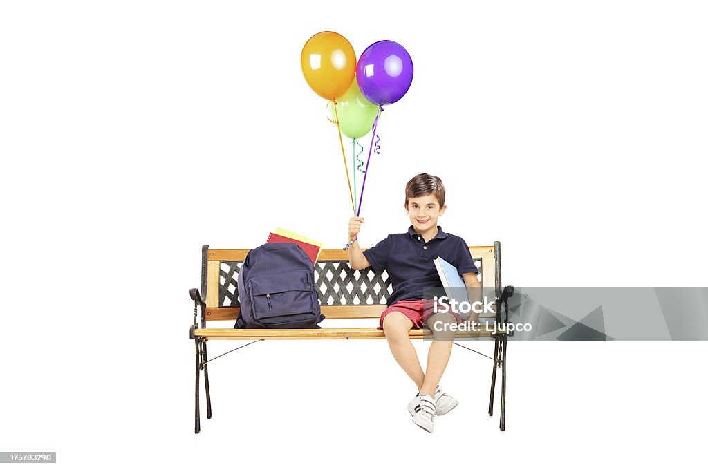 Schoolboy on bench with books holding balloons Schoolboy sitting on a wooden bench with books and holding a bunch of balloons isolated on white background Backpack Stock Photo