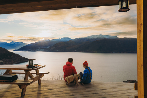 Rear View of a woman and a man in colorful jackets enjoying weekend with picnic in traditional gapahuk with a pug, contemplating a view of sea and the mountin peaks in soft light in Scandinavia