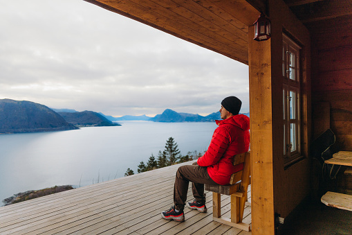 Side view of a male in red jacket sitting in the wooden shelter admiring a view of the winter sea with mountain peaks in Scandinavia