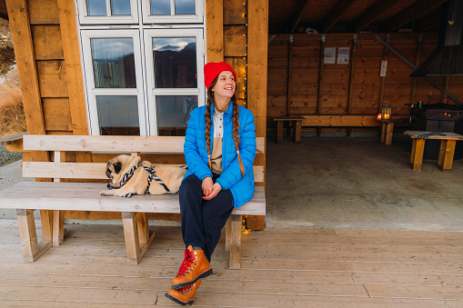 Front view of smiling female in blue jacket sitting with a dog  in the gapahuk contemplating a view of the winter sea with mountain peaks during Christmas time in Scandinavia