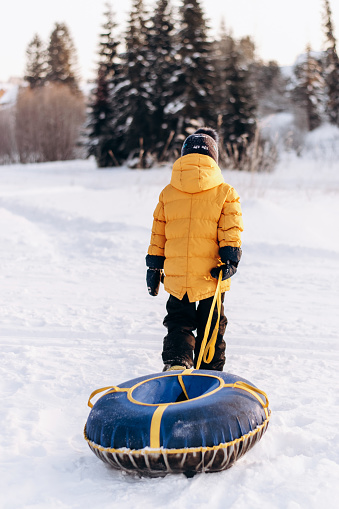 Active toddler boy in a yellow jacket pulls a rope snow tubing uphill in snow-covered forest.Winter fun,active lifestyle concept.