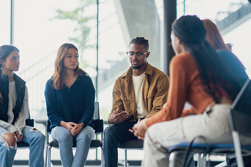 A small group of University students sit together during a group therapy session.  They are each dressed casually as they take turns sharing their struggles with the group.