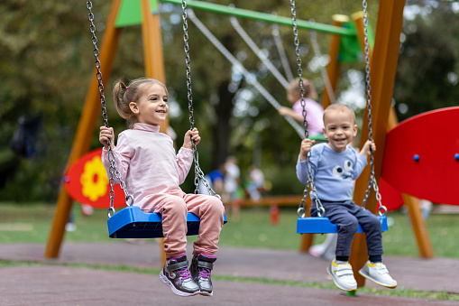 Two cute children swinging on playground outdoors.