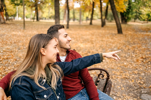 Young couple in love sitting in city park on yellow leaves in Autumn pointing with finger