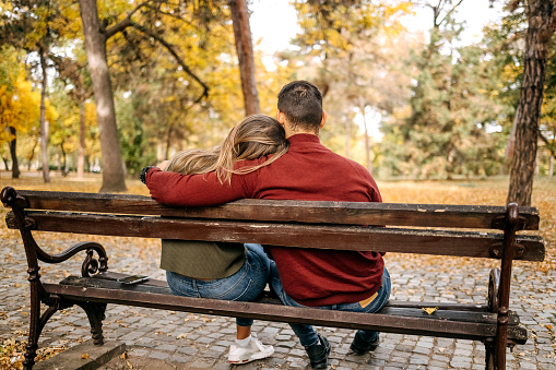 Back view of young couple in love sitting on park bench in city park on yellow leaves in Autumn