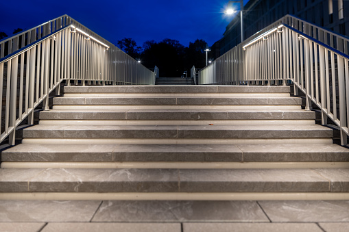 Outdoor, exterior gray stone staircase at night with chrome railings that have built in lighting.  Focus is isolated to the top stair tread and riser.