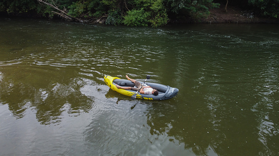 Young man with sunglasses is laying in his inflatable canoe. He is resting from rowing on the river. Photo is taken with flying drone camera.