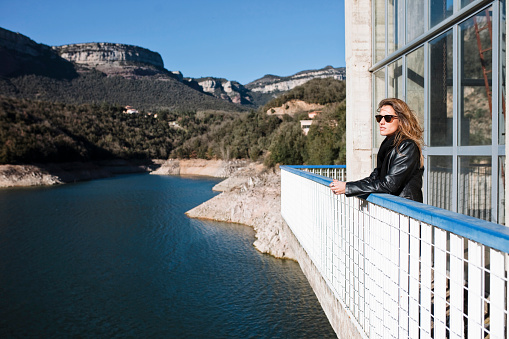 Portrait of stylish young woman, wears black jacket and eyeglasses, standing on balcony outdoors in front of lake during autumn, winter.