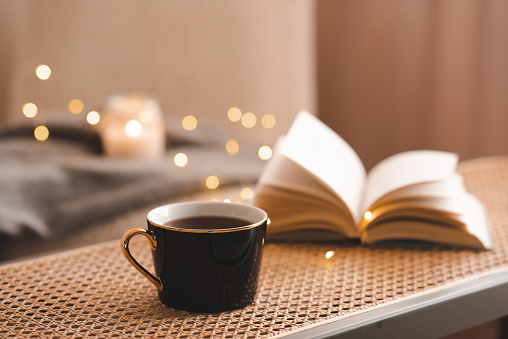 Cup of coffee with open paper book on rattan table in bedroom indoors close up over scented candle. Cozy and hygge atmosphere. Aromatherapy. Apartment living. Winter holidays