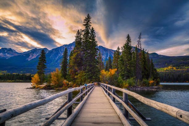 Pyramid Lake with the wooden bridge leading to Pyramid Island in Jasper National Park, Alberta, Canada. The blue sky reflects in the calm waters, framed by evergreen forests and rugged peaks of the Canadian Rockies. A breathtaking and iconic Canada travel destination perfect for themes of nature, adventure, exploration, and scenic landscapes.