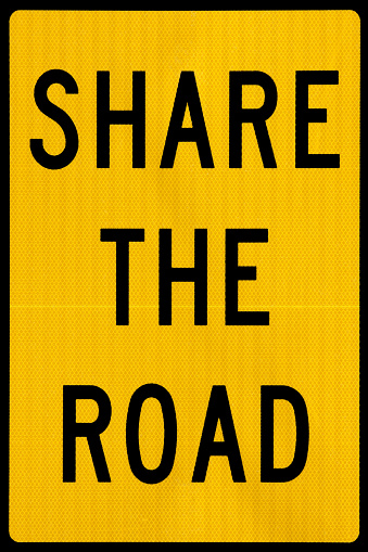 Road sign cautioning motorists to remain alert for cyclists, pedestrians, ranchers and herders who might also by using the road.  Full-frame tight crop on the sign - no hardware, posts or background appearing.  Two colors - black text and border on amber-yellow reflective backdrop.