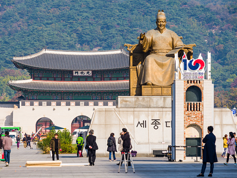 Tourists and locals strolling through the Gwanghwamun Plaza overlooked by the statue of King Sejong and the green slopes of Bukhansan mountain beyond, Seoul, South Korea.