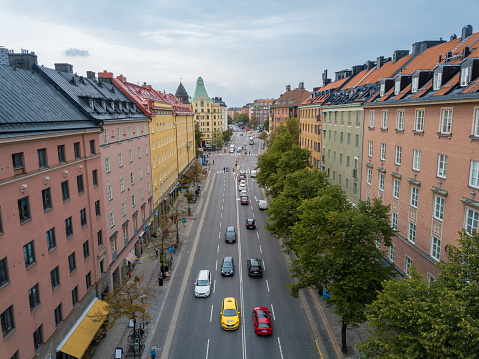 This image, taken from a drone's vantage point, beautifully captures a street gracefully winding its way through a quintessential residential area of Stockholm. The houses, exhibiting traditional Scandinavian architecture, feature vibrant facades and sharp-angled roofs. Trees, some perhaps centuries old, line the pathways, their canopies casting dappled shadows onto the roads below. In the distance, other notable Stockholm landmarks subtly rise above the residential skyline, offering a hint at the city's expansive urban layout. The overhead perspective provides a unique insight into the organized yet cozy nature of Stockholm's neighborhoods.