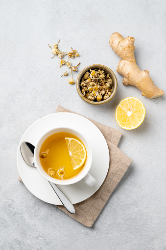 Chamomile herbal tea with lemon in a white cup on a light background with dry flowers and ginger. The concept of a healthy detox drink for health and immunity. Top view.