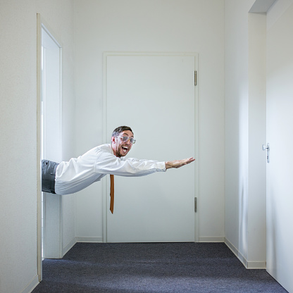 A funny stereotypical office worker has the ability to fly, and he smiles big at the camera as he is flying through an office hallway.  Square crop with copy space.