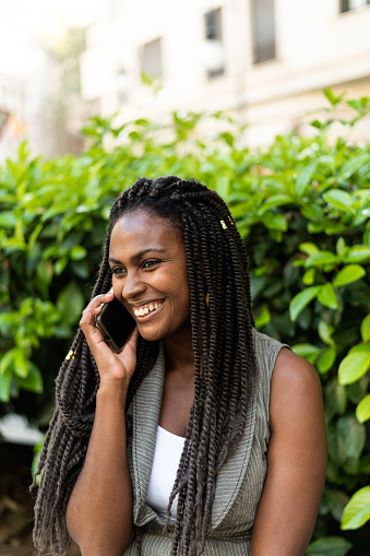 Vertical photo with copy space of smiling african woman with braided hair talking to the mobile on an urban park