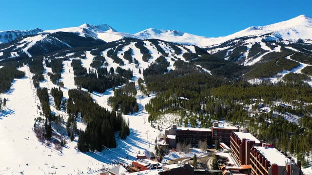 Aerial Drone View of Giant Ski Resort with People Skiing by Full Parking Lot and Riding Chairlifts Down Mountain Slopes through a Pine Tree Forest Covered in Snow