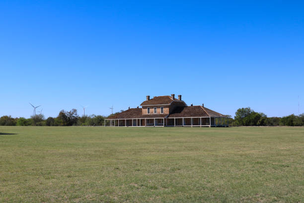 view of the hospital at Ft. Richardson, a long porch across the entire front. with windows and a single door in the middle of the building, a small upper floor with 3 windows and 4 chimneys, backed by a bright blue clear sky in the autumn weather, surrounded by trees in the back and grass in the front, wind vanes in the background