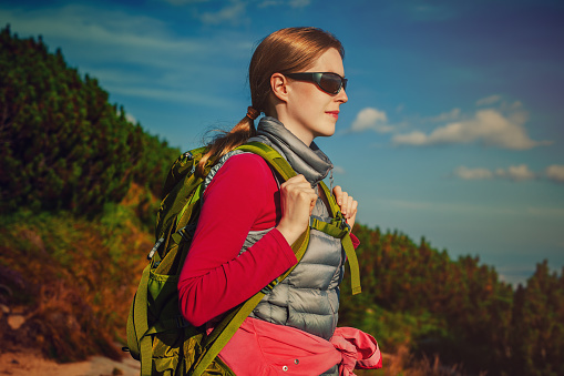Young woman tourist with green backpack and sunglasses standing on mountains background