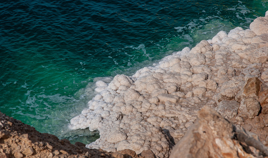 Dead Sea coastline in Jordan. Beautiful cliffs cascade down into emerald water of Dead Sea. Salt crystals show in strips level of leaving water.  Ecology theme