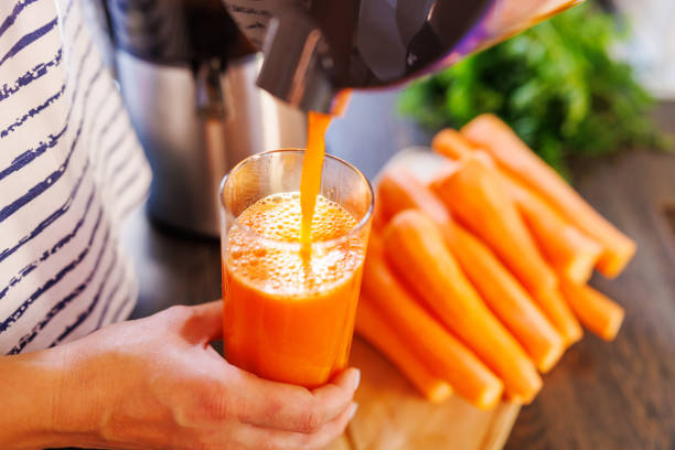una mujer vierte jugo de zanahoria fresco en un vaso. zumo de zanahoria recién exprimido - freshly squeezed fotografías e imágenes de stock