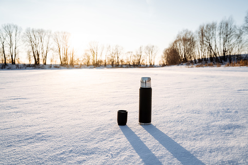 A thermos of black with a cup stands on the snow. Tourist utensils. Drink hot tea in winter to keep warm. Vacuum bottle for hot water. Glare of the sun in nature. High quality photo