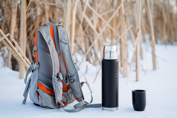 istock A black thermos of tea stands next to the backpack. Vessel for hot drinks. A small backpack for a hike. Rest on the lake. 1758290620