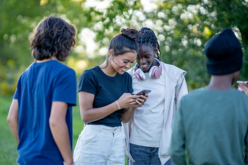 A small group of teenagers stand around outside on a warm summers day, with their cell phones in hand, as they connect online and laugh with one another.