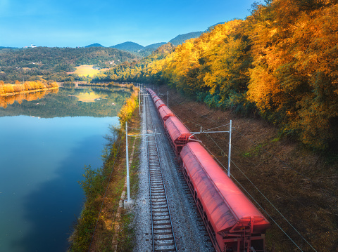 Aerial view of red freight train moving near river in alpine mountains at sunrise in autumn. Top view of wagons, railroad, lake, reflection in water, orange trees in fall. Railway station in Slovenia